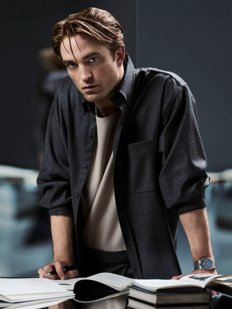 Robert Pattinson leaning over a desk of open notebooks with a pen in hand, wearing an overshirt over a crewneck and a steel Jaeger-LeCoultre watch