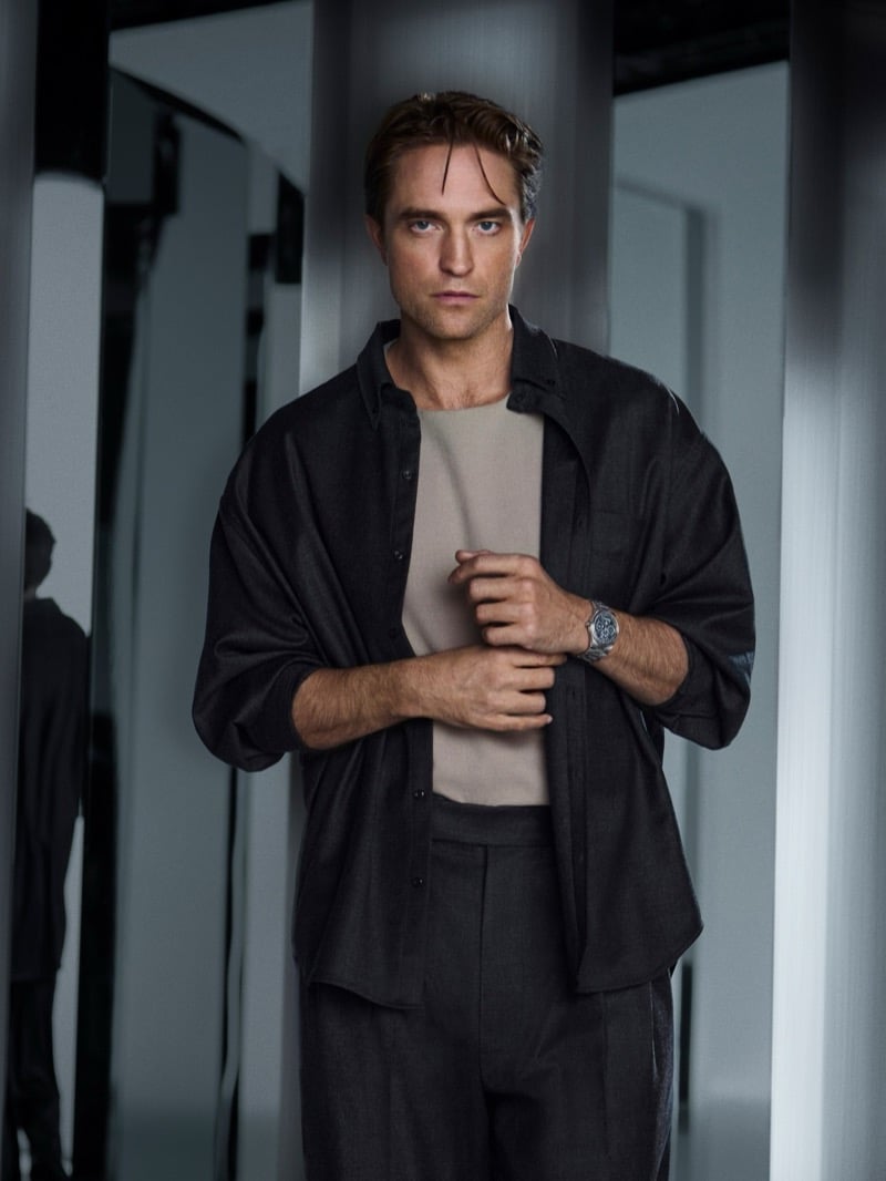 Robert Pattinson standing among mirrored panels in an open overshirt and pleated trousers, adjusting his steel Jaeger-LeCoultre watch