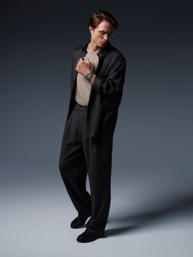 Robert Pattinson standing in an open overshirt and wide pleated trousers, clasping his collar with a Jaeger-LeCoultre watch on his wrist