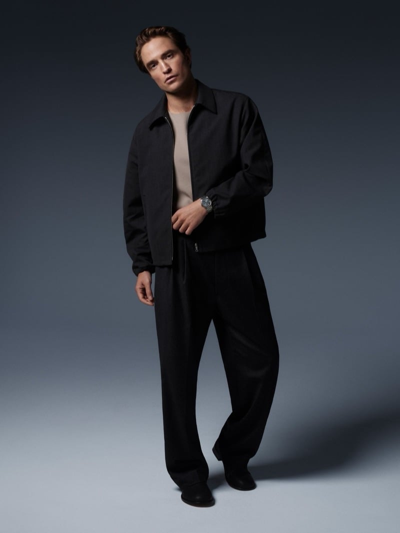 Full-length view of Robert Pattinson in a zip-front jacket, crewneck, and wide pleated trousers wearing a Jaeger-LeCoultre watch