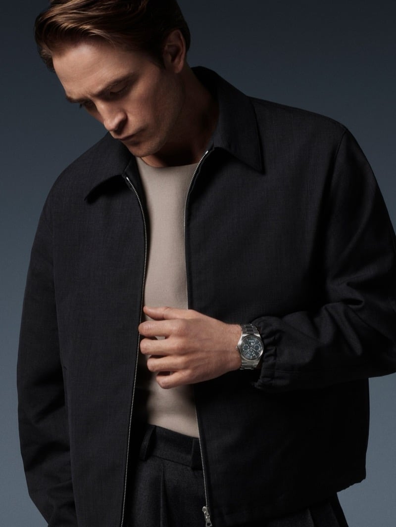 Robert Pattinson looking down as he holds the front of a zip jacket, a Jaeger-LeCoultre watch at the cuff