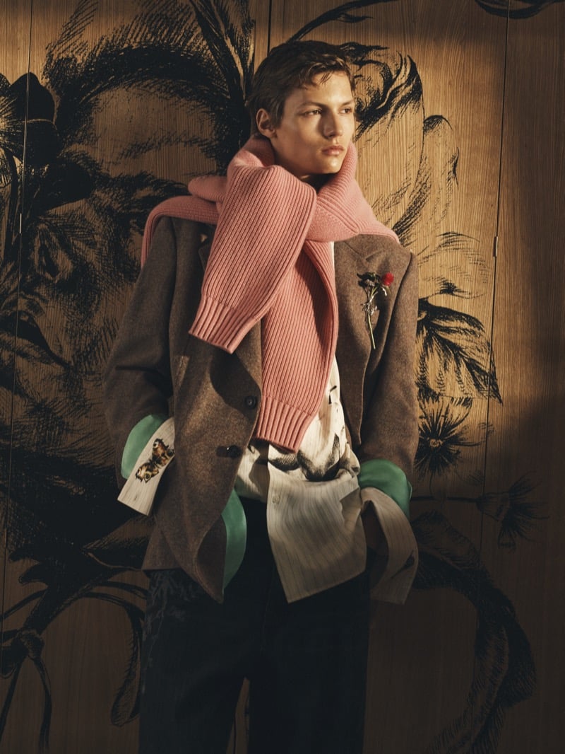 Roman Lenoir in a brown wool blazer, pink sweater draped over his shoulders, a printed shirt with butterfly cuffs against a painted wooden wall