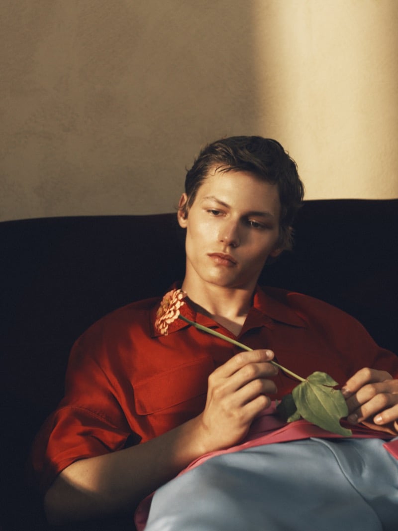 Roman Lenoir seated on a sofa in a red short-sleeve shirt, holding a single flower by its stem
