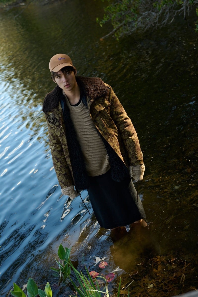 Rui Borysevicz stands barefoot in a lake wearing a Dior cap, botanical-print jacket with a shearling collar, ribbed sweater, and long knit skirt