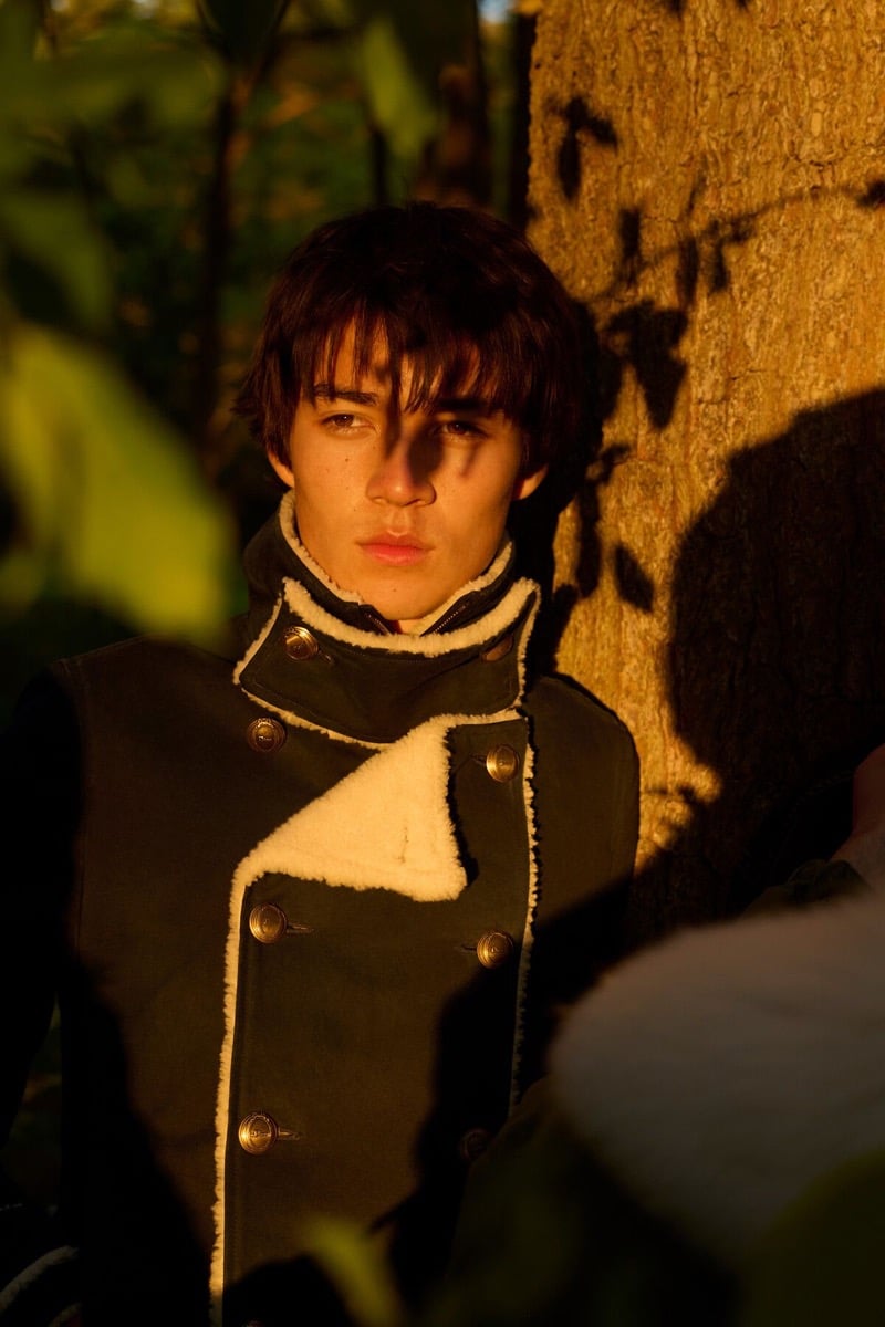 Rui Borysevicz in a double-breasted Dior coat with shearling trim and brass buttons leans against a tree in golden-hour light