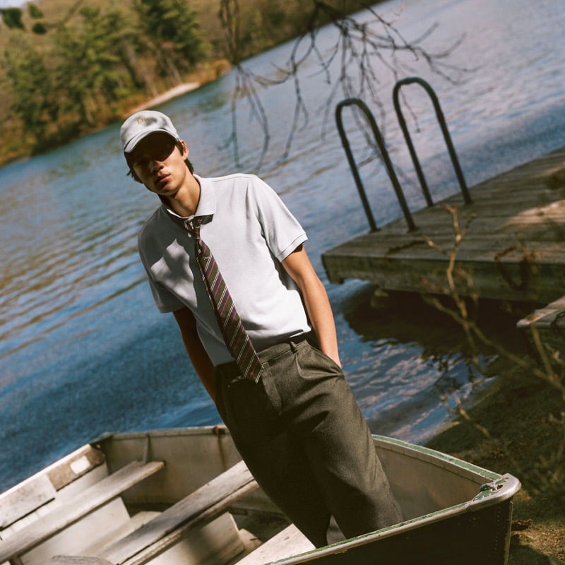 Rui Borysevicz in a Dior cap, short-sleeve polo, striped tie, and wool trousers steps into a rowboat beside a lake dock