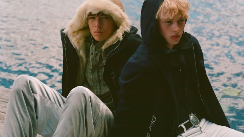 Rui Borysevicz in a shearling-trimmed hood and checked shirt sits on a dock beside Quinn McLachlan in a hooded Dior jacket and logo belt