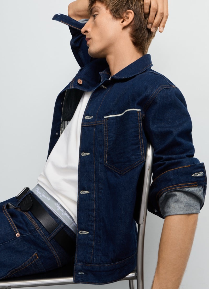 Samuel Curtis sits with a hand through his hair in Pepe Jeans' selvedge trucker jacket over the branded Dalston jean waistband