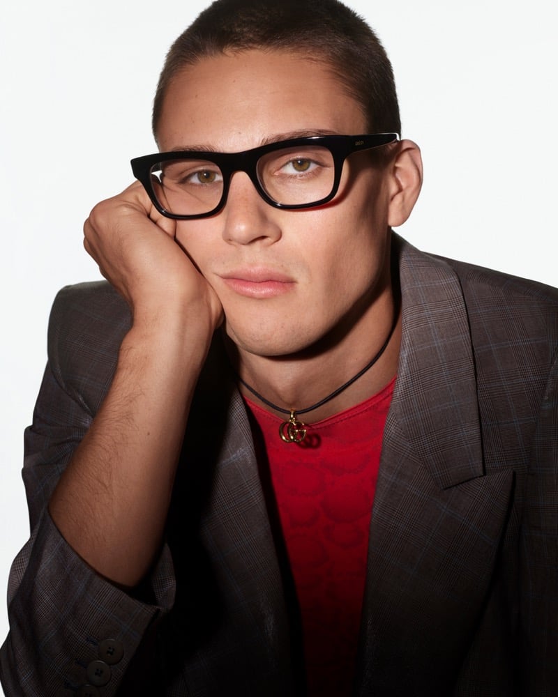 Samuel Watson wears black rectangular Gucci optical glasses and a gray check blazer over a red top with a Double G pendant necklace for Gucci Fall-Winter 2026 eyewear.