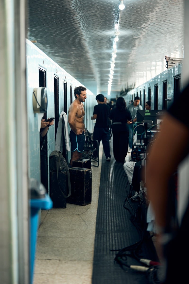 Sebastián Zurita stands shirtless backstage in a locker room hallway while crew members work around him, for Original Penguin's fall 2026 campaign