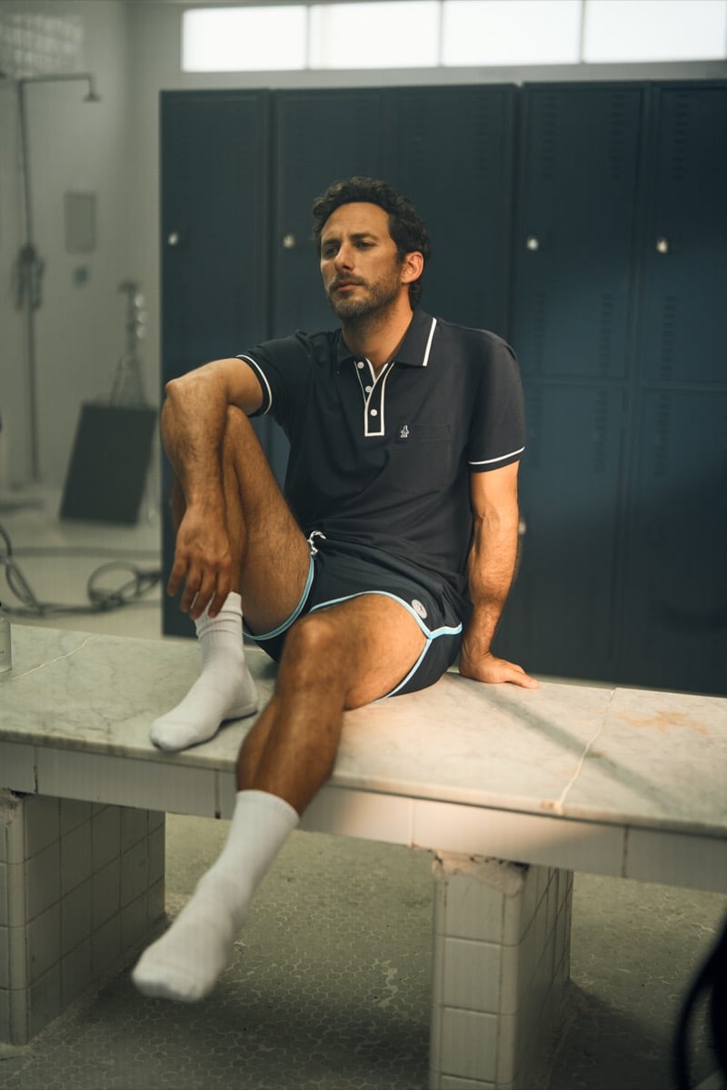 Sebastián Zurita sits on a marble locker room bench in a white-trimmed polo and teal-piped shorts, for Original Penguin's fall 2026 campaign