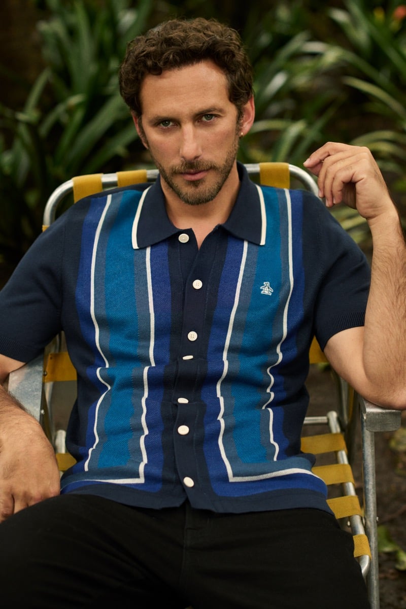 Sebastián Zurita rests an arm on a folding garden chair in a blue-striped knit polo and black trousers, for Original Penguin's fall 2026 campaign