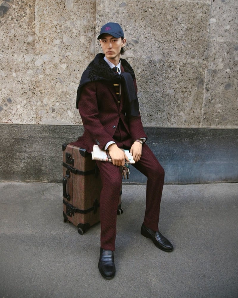 Simon Martyn sits on a paisley-printed hard-shell suitcase in a burgundy textured suit, white shirt, and dark scarf, holding a folded newspaper and keys, wearing a dark Etro cap, for the Etro fall 2026 campaign