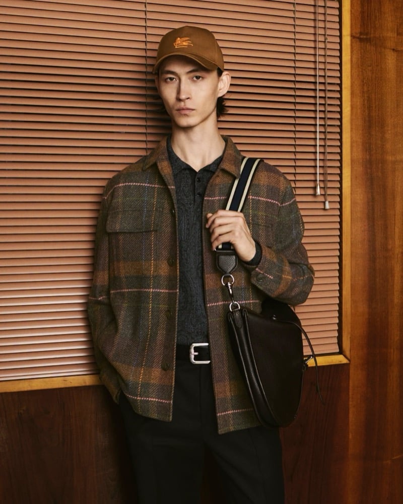 Simon Martyn stands in front of wooden window blinds in a brown and green plaid wool overshirt over a dark knit polo with dark trousers, a brown Etro logo cap, and a black leather crossbody bag on a striped strap, for the Etro fall 2026 campaign