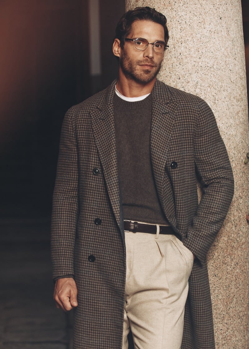 Simone Bredariol against a stone column in a brown houndstooth overcoat over a sweater with pleated trousers, for Boglioli's fall-winter 2026 campaign