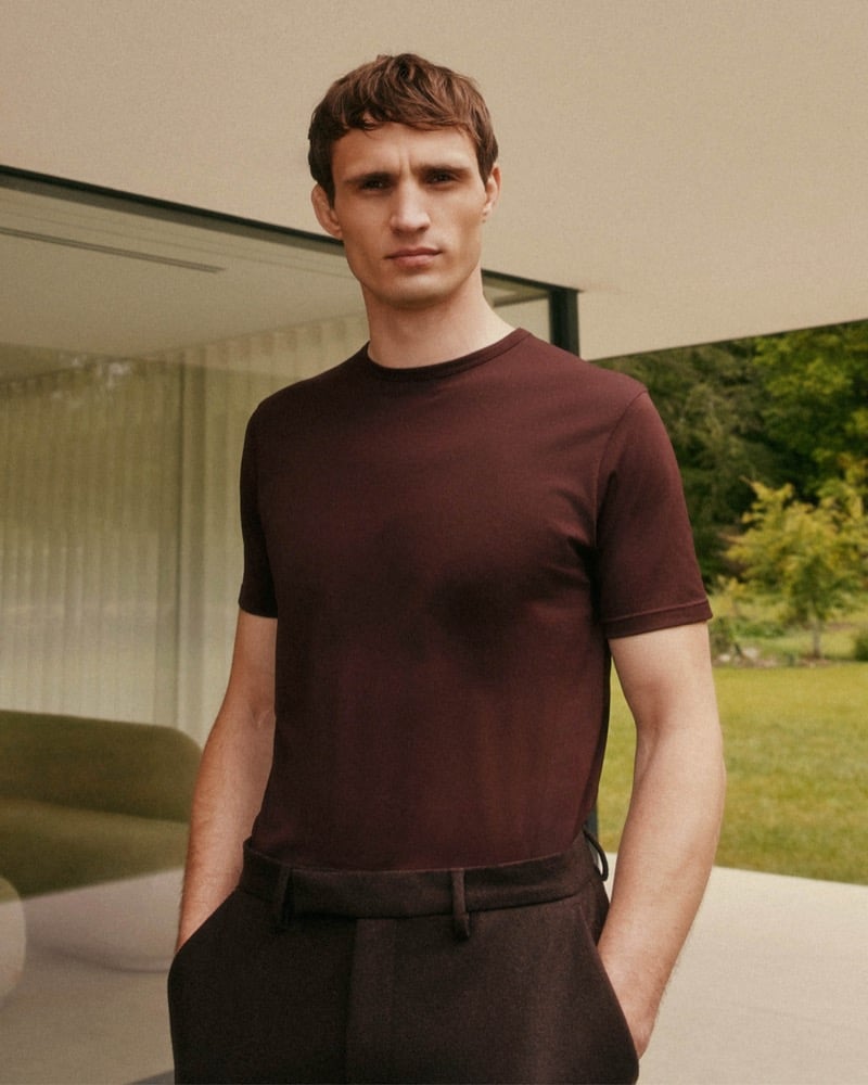 Julian Schneyder stands on a terrace outside a glass pavilion, wearing a burgundy short-sleeve cotton T-shirt tucked into brown tailored trousers