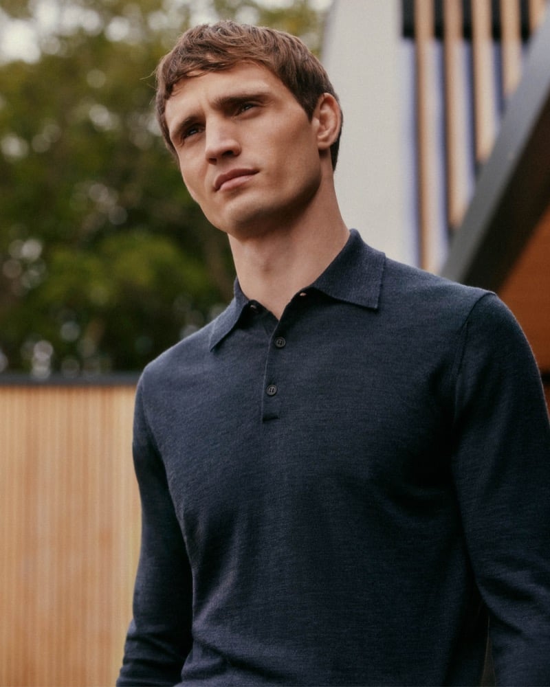 Julian Schneyder stands outdoors against greenery and a slatted wood wall, wearing a dark merino long-sleeve knit polo shirt