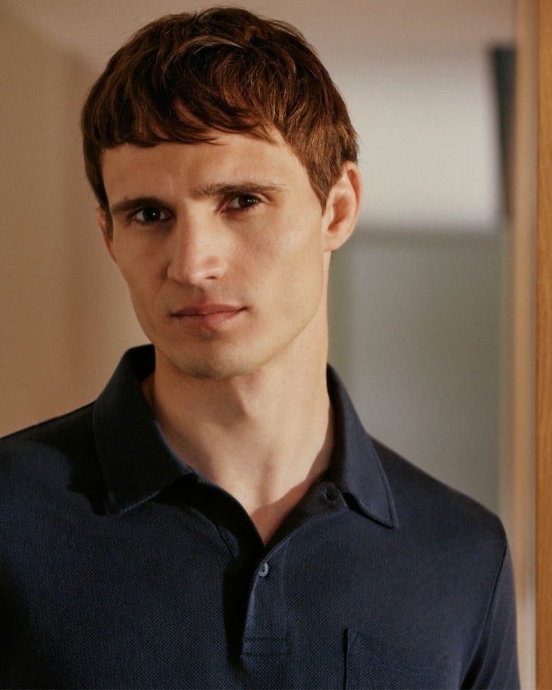 Close portrait of Julian Schneyder in a warmly lit hallway, wearing a navy textured pique polo shirt with the collar open