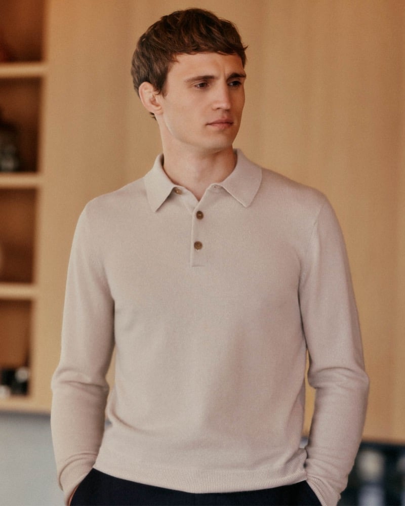 Julian Schneyder stands in front of a pale wood cabinet with his hands in his pockets, wearing an oatmeal long-sleeve knit polo with three horn buttons and dark trousers