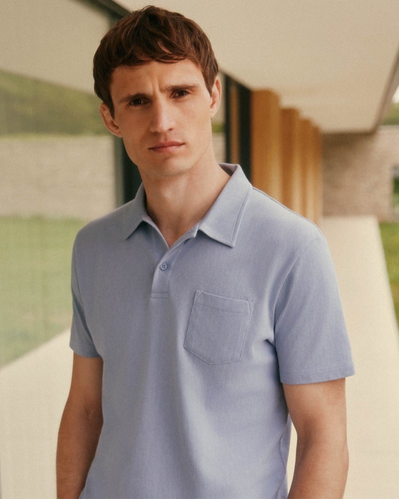 Julian Schneyder photographed under a concrete overhang, wearing a powder-blue pique short-sleeve polo shirt with a chest pocket