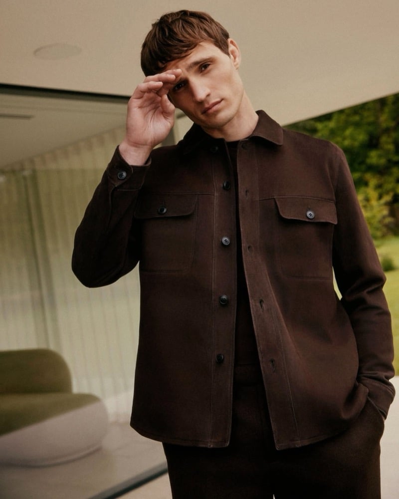 Julian Schneyder stands in a glass-walled room with one hand raised to his brow, wearing an open brown suede overshirt with two chest pockets over a brown crew-neck knit and matching trousers