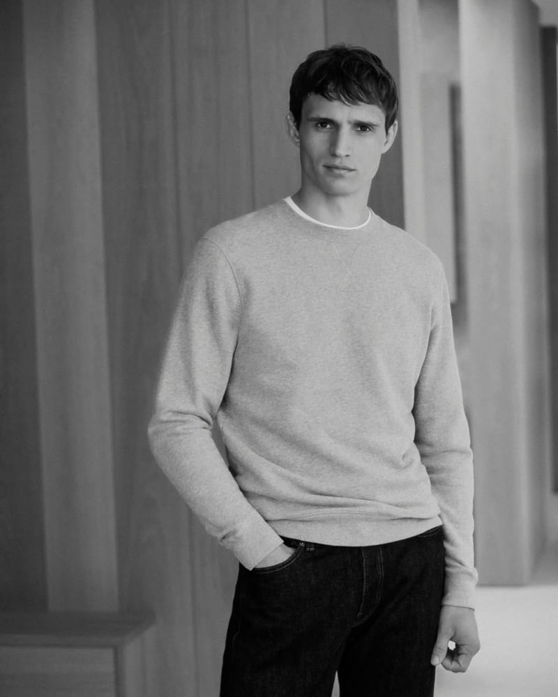 Black-and-white portrait of Julian Schneyder standing in a wood-panelled interior with one hand in his pocket, wearing a loopback crew-neck sweatshirt over a T-shirt with dark denim jeans