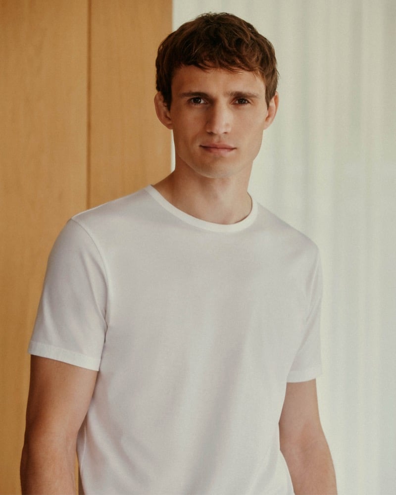 Julian Schneyder in front of a pale wood panel and a sheer curtain, wearing a crisp white cotton crew-neck T-shirt