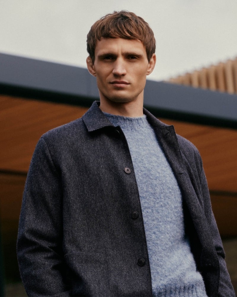 Julian Schneyder photographed outdoors in front of a timber building, wearing an unbuttoned flecked blue wool overshirt over a brushed powder-blue crew-neck sweater