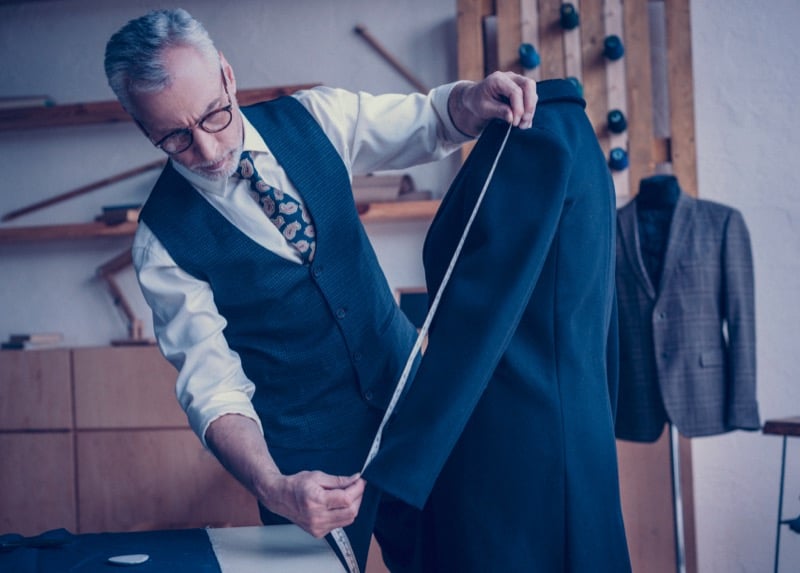 An older tailor in a waistcoat and tie measures the length of a dark overcoat with a tape measure in his workshop