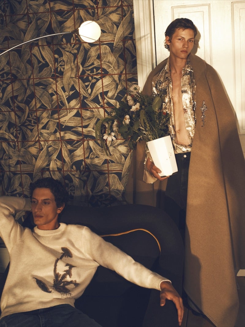 Takfarines Bengana seated in a sweater with a dandelion motif as Roman Lenoir stands in a sequin shirt and draped camel coat holding a vase of flowers
