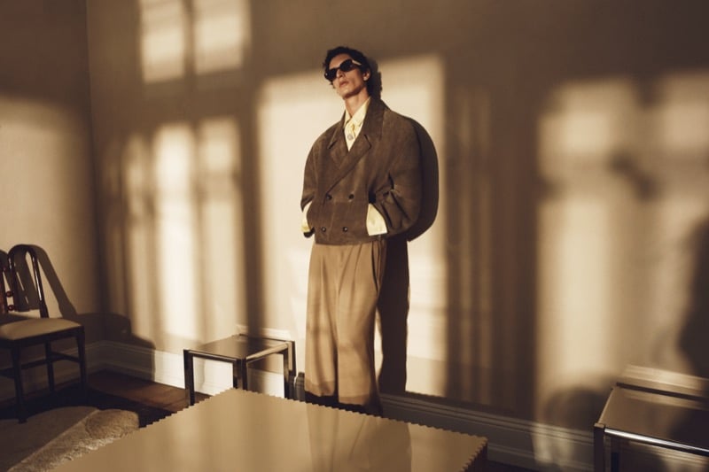 Takfarines Bengana in sunglasses, an oversized double-breasted suede jacket, and wide pleated trousers standing in a sunlit room