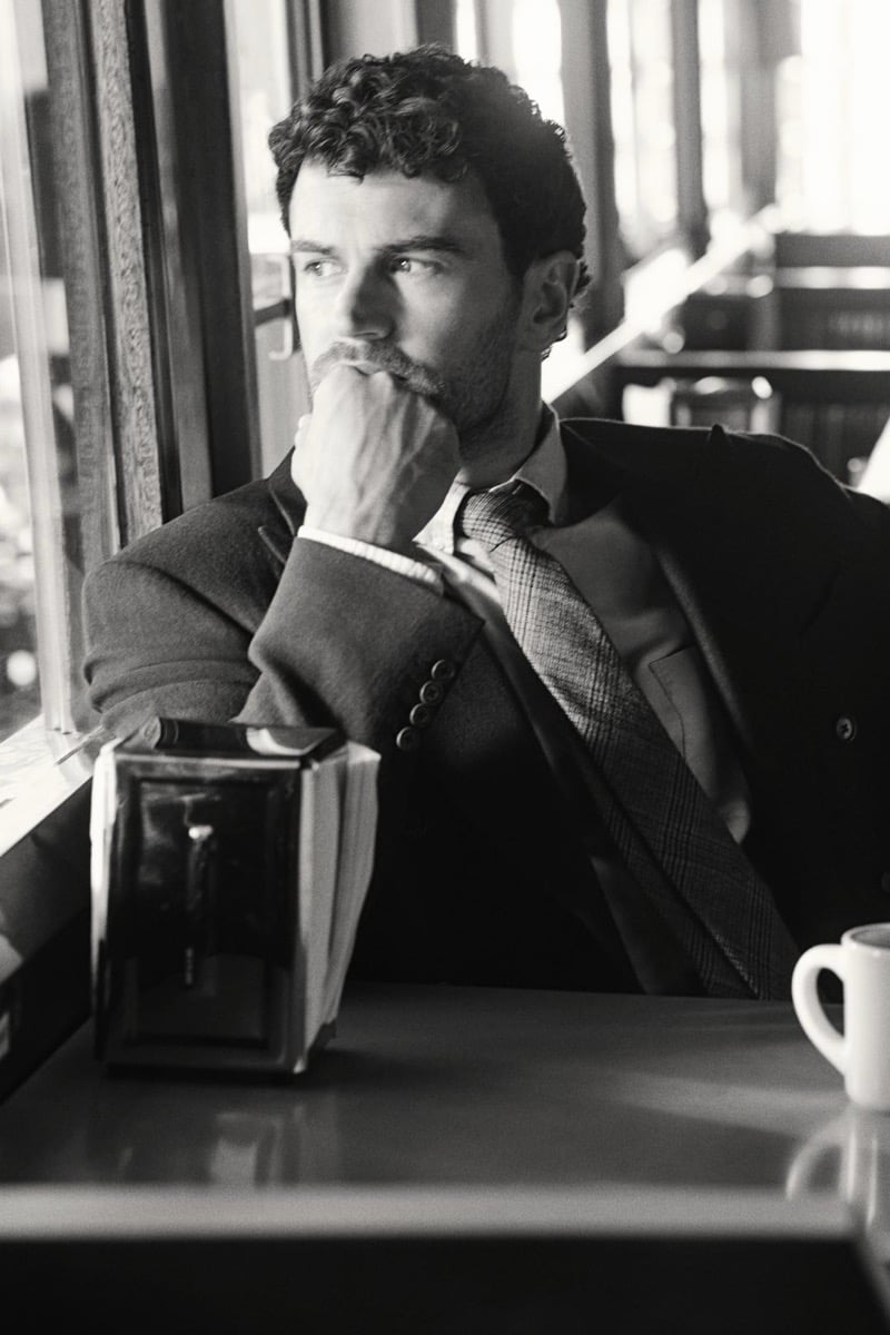 Black and white shot of Theo James seated at a diner window with a coffee cup on the table, resting his chin on his hand, wearing a double-breasted blazer over a shirt and check tie