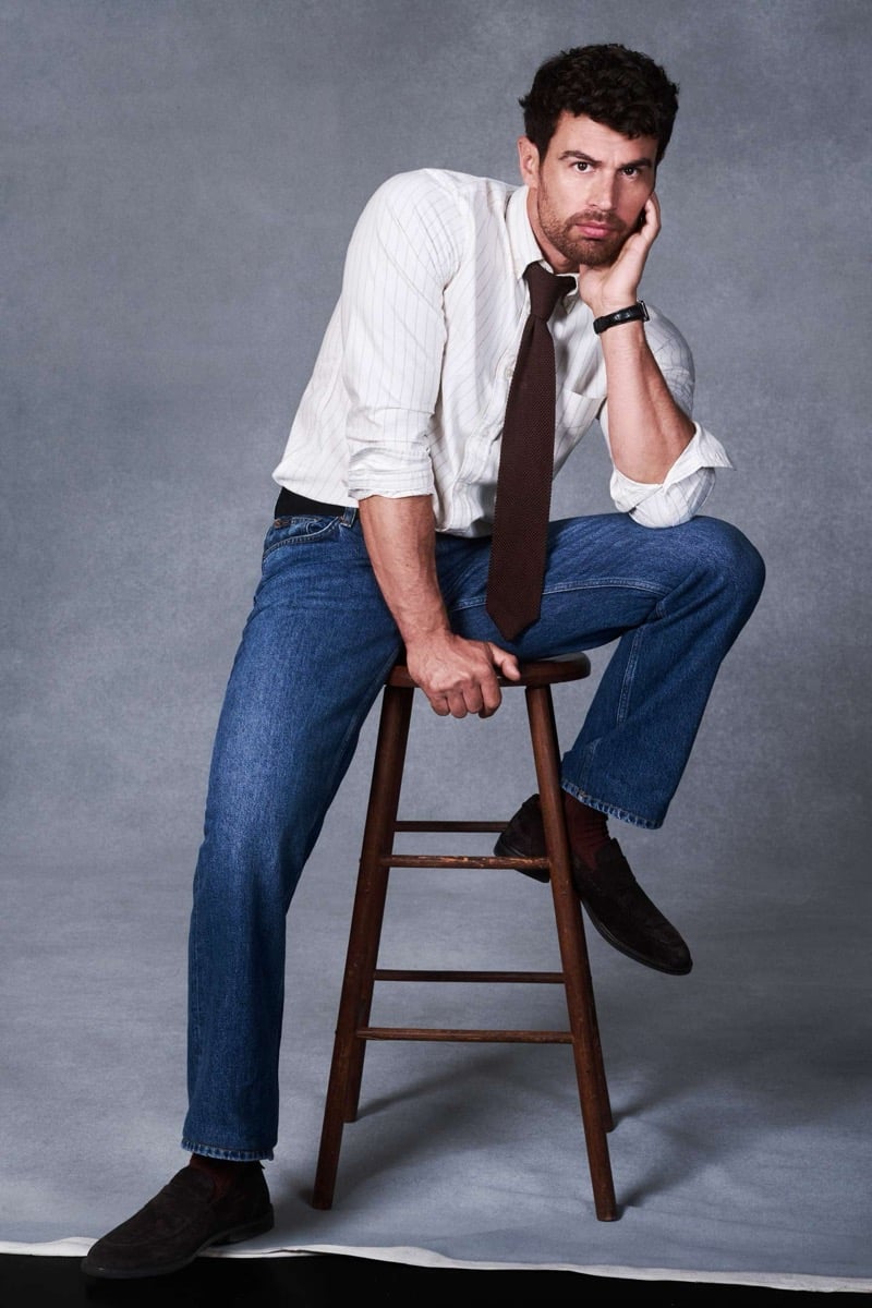 Theo James sits on a wooden stool against a grey studio backdrop, wearing a striped Oxford shirt with the sleeves pushed up and a loosened brown knit tie, mid-wash straight-leg jeans, and brown suede loafers