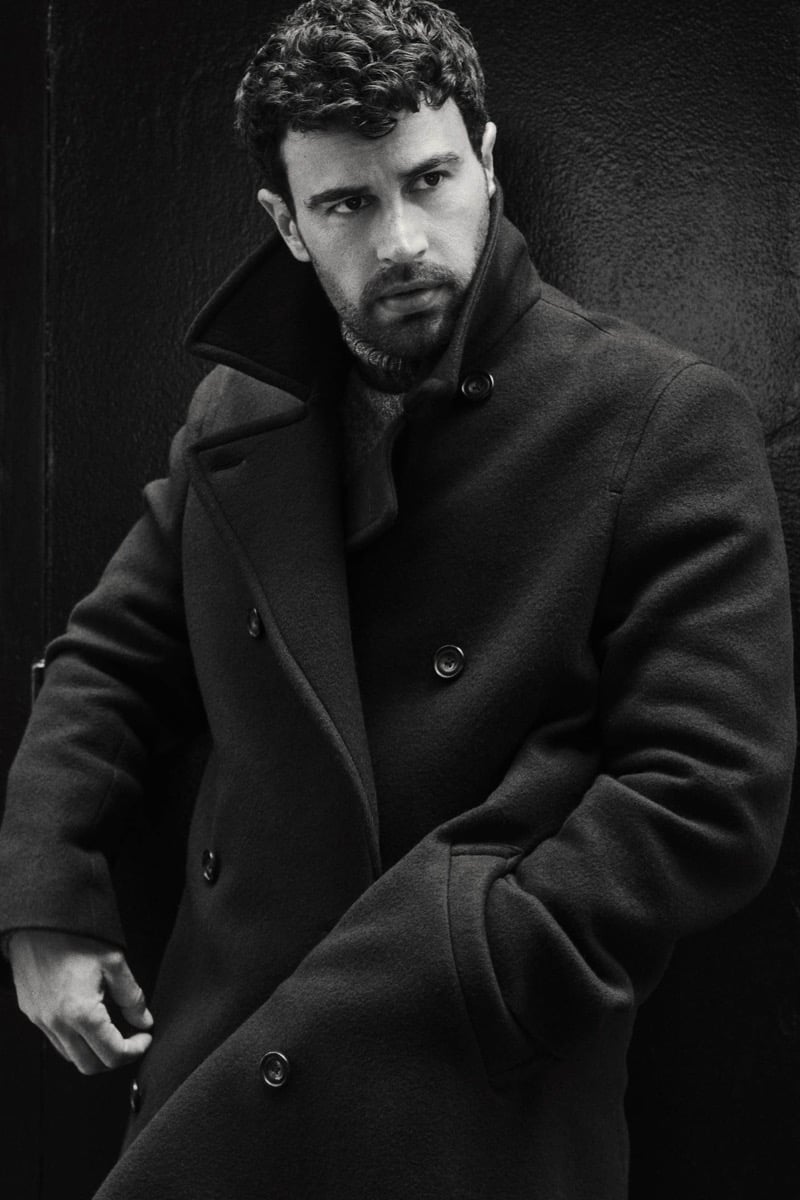 Black and white close-up of Theo James with the collar of a double-breasted wool overcoat turned up over a turtleneck, against a dark wall