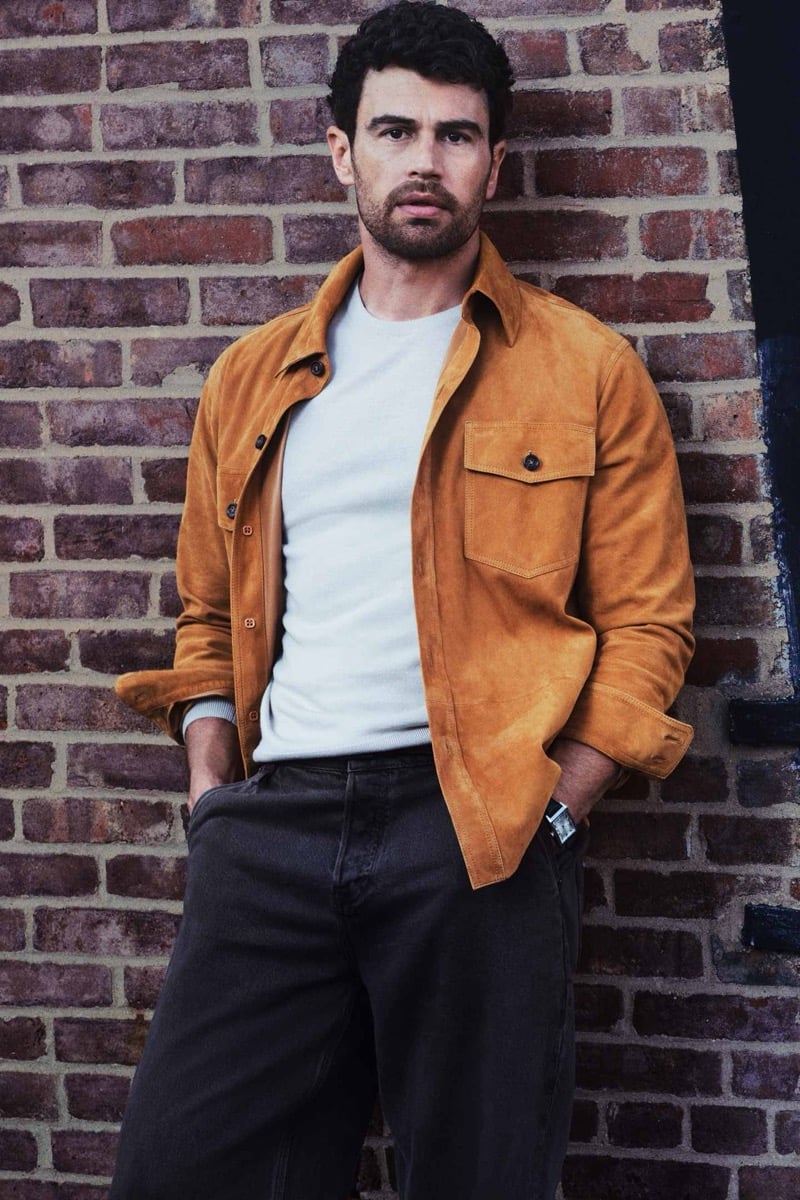 Theo James stands against a red brick wall with his hands in his pockets, wearing an unbuttoned tan suede overshirt over a light crew-neck knit with dark jeans and a wristwatch