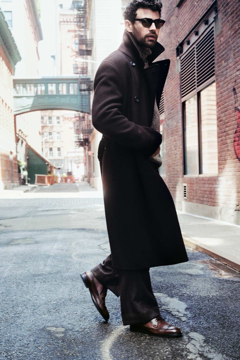 Theo James walks across a cobbled Tribeca street in sunglasses and a long brown wool overcoat worn open over a knit turtleneck, with dark trousers and brown brogues