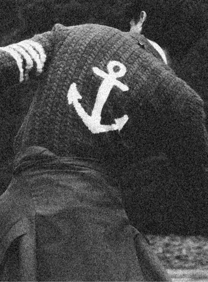 Grainy black-and-white close-up of the back of a chunky Thom Browne hand-knit sweater with a large anchor motif and a four-bar stripe on the sleeve