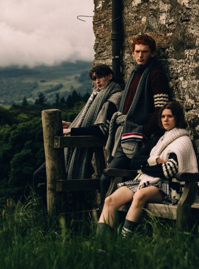Arthur del Beato, Arran Young, and a female model sit close together on a weathered bench by a stone ruin, layered in Thom Browne cable-knit sweaters, scarves, and a check skirt