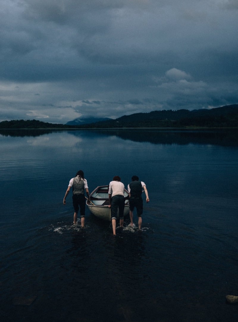 Three models wade into a still loch at dusk pushing a small boat, barefoot with trousers rolled, in Thom Browne white shirts and waistcoats, mountains reflected in the water