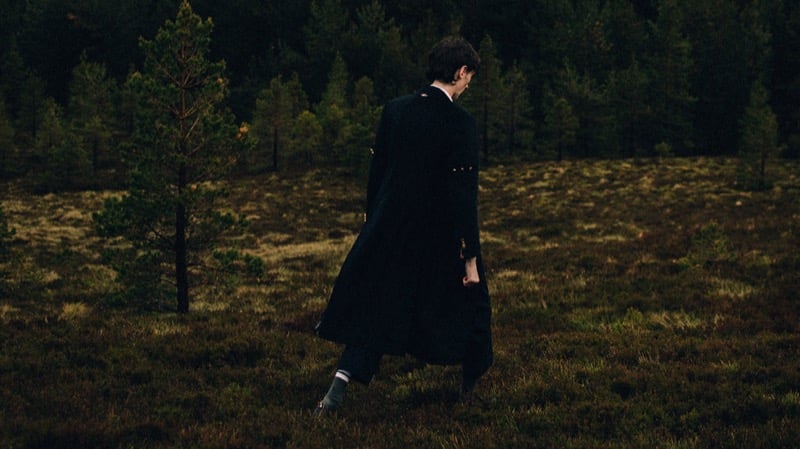 Arran Young walks away across open moorland toward a pine wood, wearing a long dark Thom Browne brass-button coat and striped socks