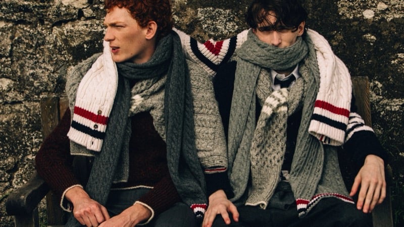 Arthur del Beato and Arran Young sit against a stone wall wrapped in oversized Thom Browne cable-knit sweaters and chunky gray scarves, one in a burgundy marled knit, the other in a striped V-neck sweater and tie