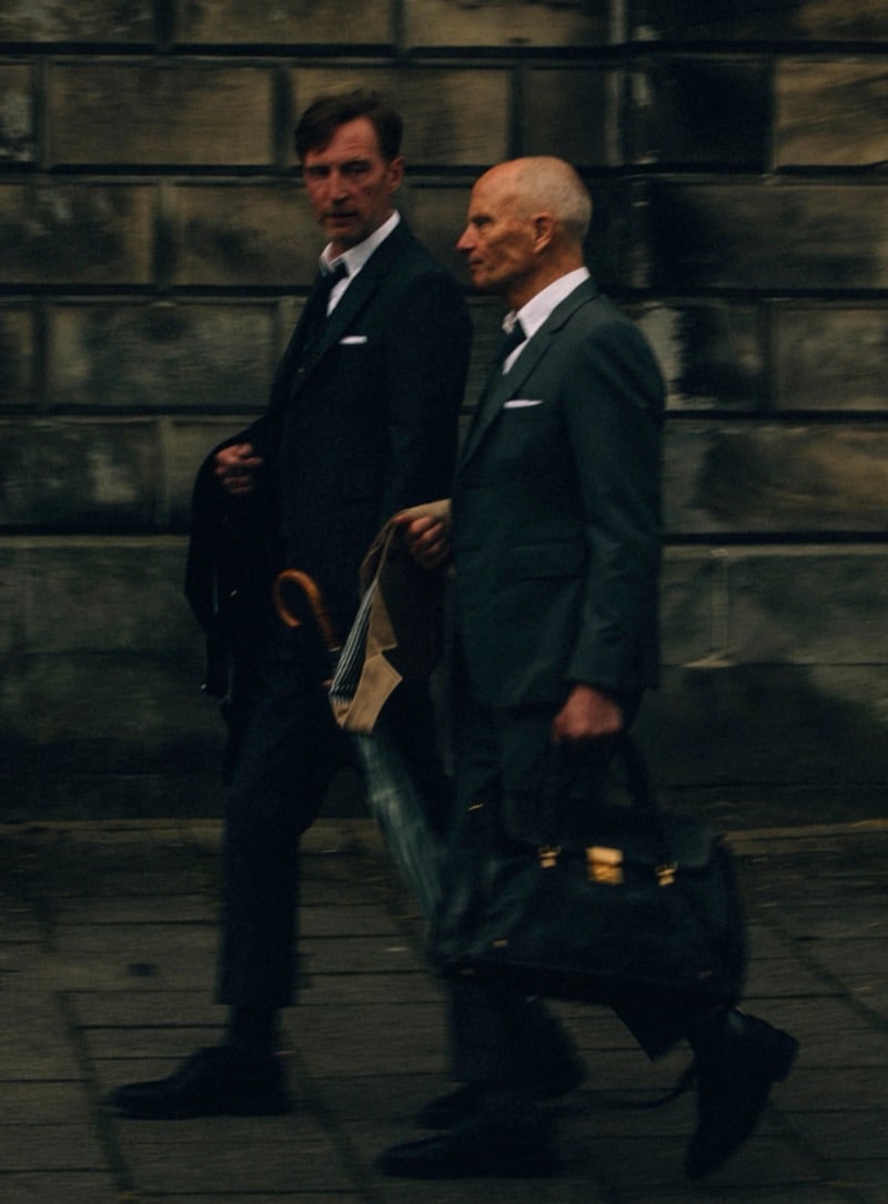 Two older models walk past a stone wall in tailored Thom Browne suits with ties and white pocket squares, one carrying a leather doctor's bag and a striped umbrella