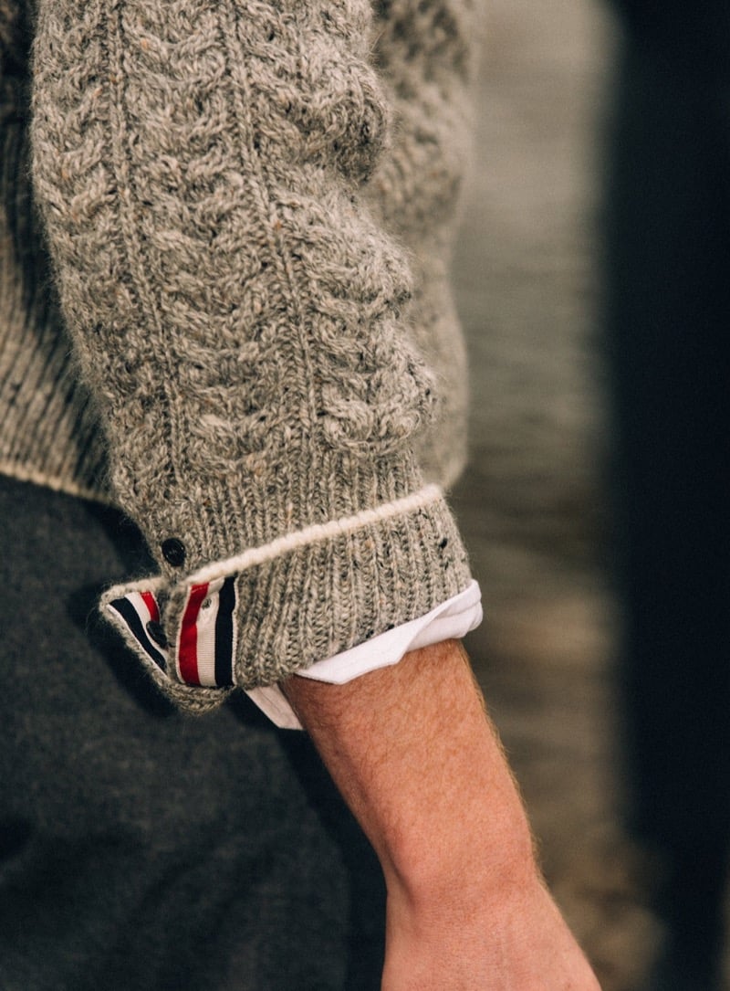 Close-up of a gray Thom Browne cable-knit sweater cuff with a buttoned tab lined in red, white, and blue grosgrain, folded back over a white shirt cuff