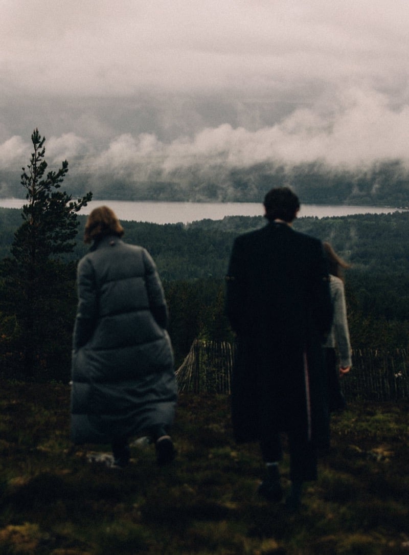 Two figures seen from behind on a ridge above a misty loch, one in a long gray Thom Browne puffer coat, the other in a long dark tailored coat