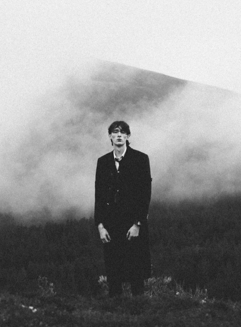 Black-and-white photo of Arran Young standing alone in heavy mist on a hillside, wearing a dark Thom Browne tailored jacket, shirt, and tie