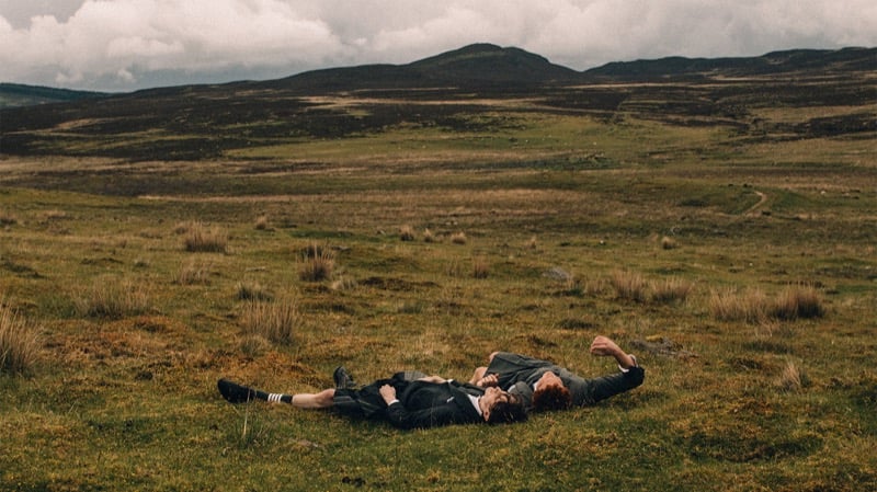 Arran Young and Arthur del Beato lie on their backs in moorland grass in Thom Browne tailoring, dwarfed by the rolling hills and heavy sky behind them