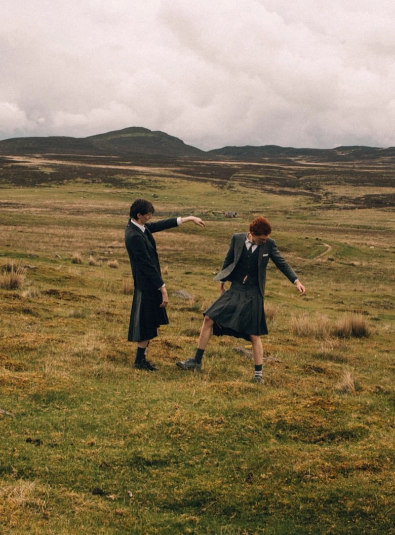 Arran Young and Arthur del Beato walk across moorland hills wearing Thom Browne tailored jackets, waistcoats, ties, and knife-pleated gray skirts with dress socks and lace-up shoes