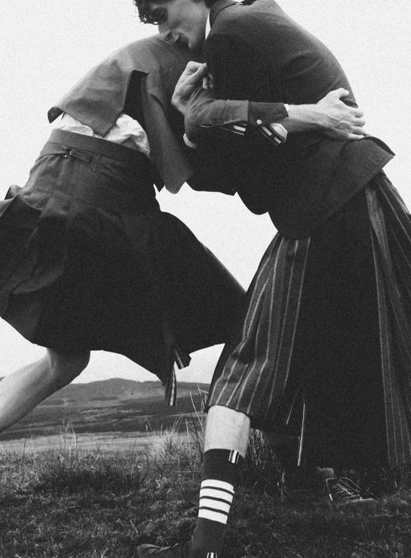 Black-and-white photo of Arthur del Beato and Arran Young wrestling on a hilltop in Thom Browne tailored jackets and pleated skirts, one in a pinstriped skirt with four-bar striped socks