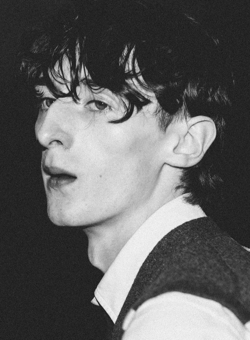 Black-and-white close-up profile of Arran Young with tousled hair, wearing a Thom Browne V-neck sweater over a white shirt collar