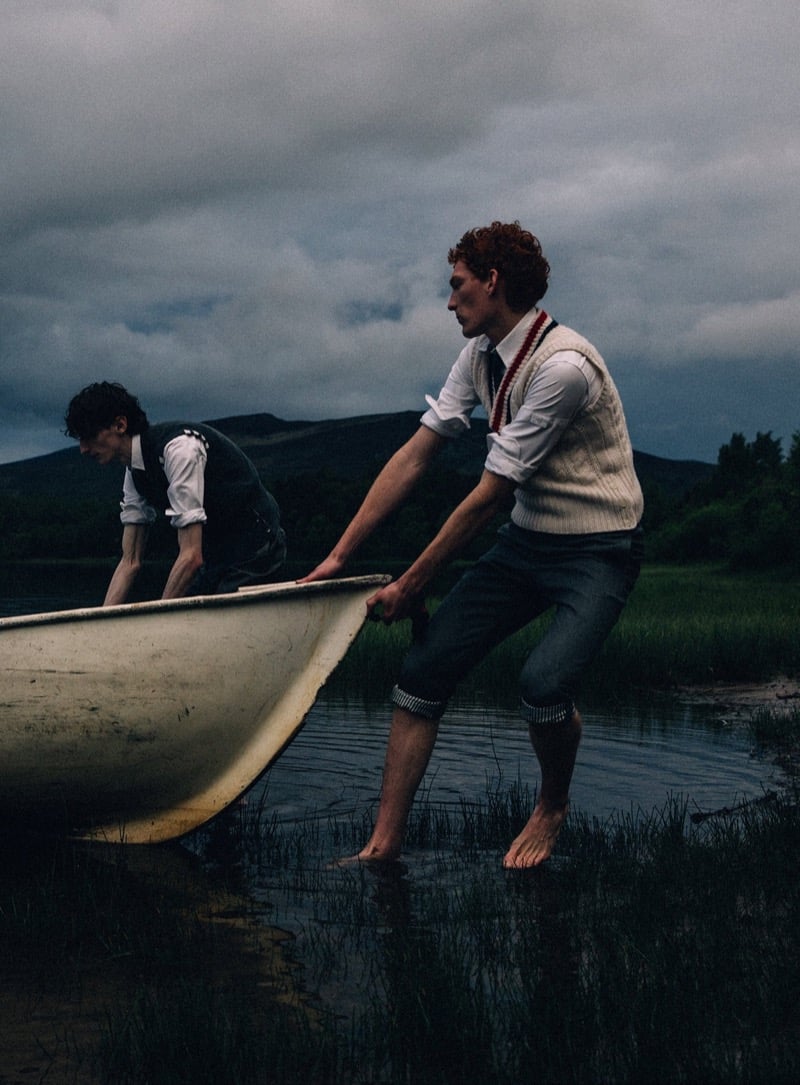 Arran Young and Arthur del Beato drag a small wooden rowboat into a dark lake at dusk, barefoot with trousers rolled up, wearing Thom Browne white shirts, a waistcoat, and a cable-knit sweater vest with a long striped knit tie